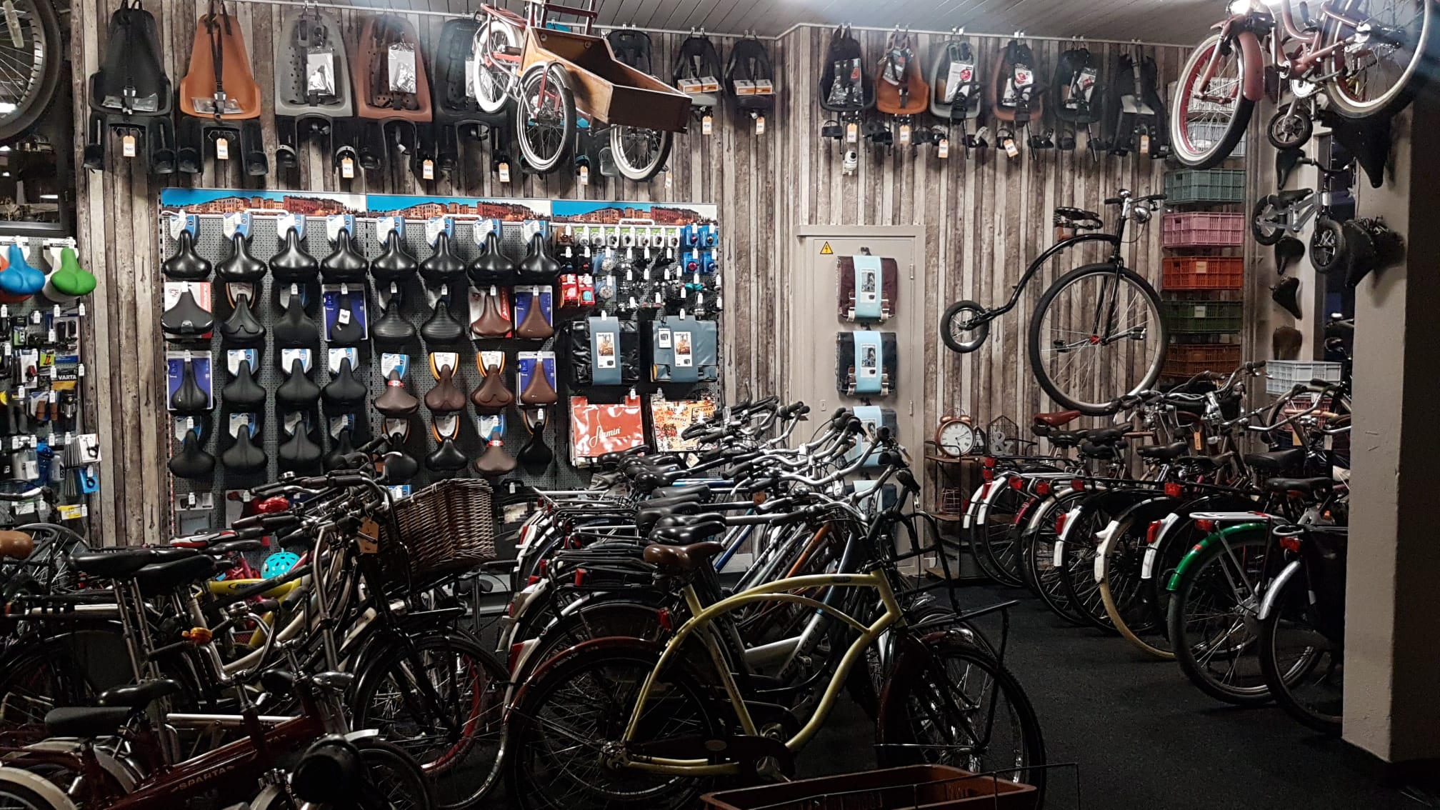 De winkel van binnen: een houten wand vol zadels, fietsstoeltjes en tassen, met rijen fietsen en een hoge bi aan de muur